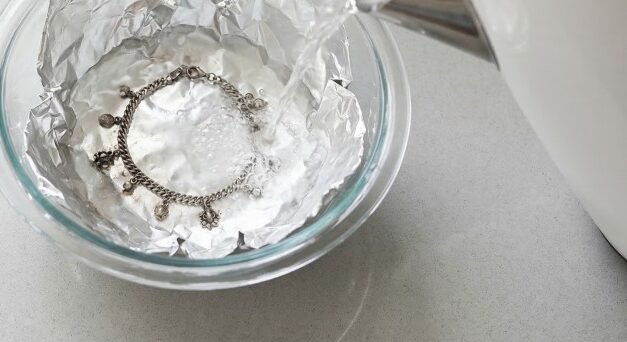 An overhead shot of a silver kettle pouring hot, steaming water into a foil-lined glass bowl containing a tarnished silver bracelet. A measuring spoon filled with baking soda rests on the grey counter.